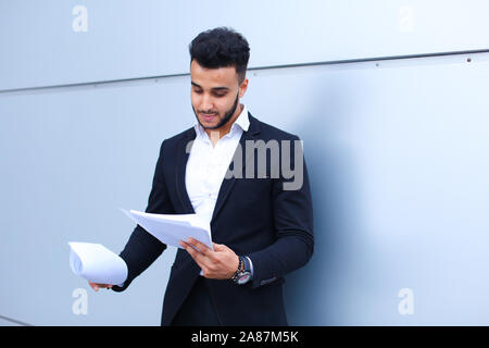 Arabian Successful businessman holds documents, smiling little smile and thinks near wall of business center. Stock Photo