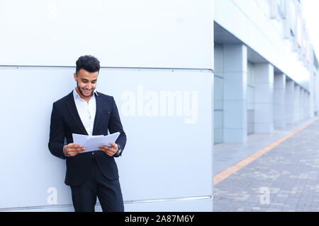 Arabian handsome student smiling, holds and looks at documents near wall of business center. Stock Photo