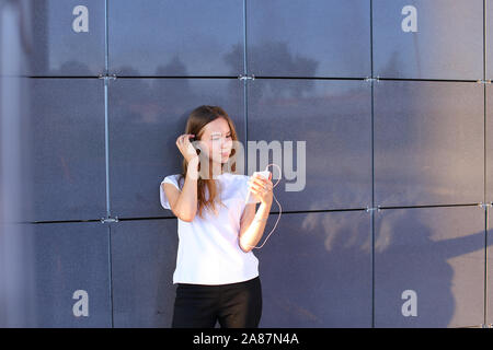 Young entrepreneur pretty female girl holds phone in hand, uses gadget, listens music, speaks on headphones rests on wall of business center. Stock Photo