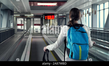 Young woman traveller on escalator with backpack and suitcase in airport terminal. She is going to to the airport exit, back view. Stock Photo