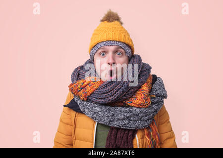 Handsome caucasian shocked funny man in several hats and scarfs being surprised on pink background. Winter fashion. Stock Photo