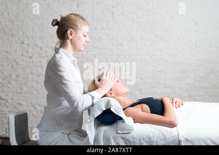 Professional female masseur giving relaxing massage treatment to young female client. Hands of masseuse on forehead of young lady during procedure of Stock Photo