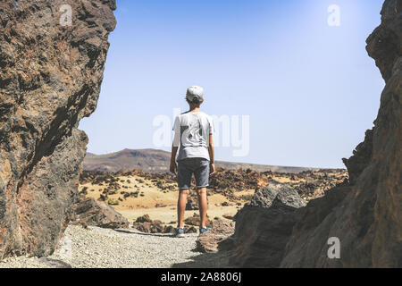 Back view of a teen, standing between lava rocks, looking at the horizon. Trendy boy looks enthusiastically at the view of the Teide National Park. Stock Photo