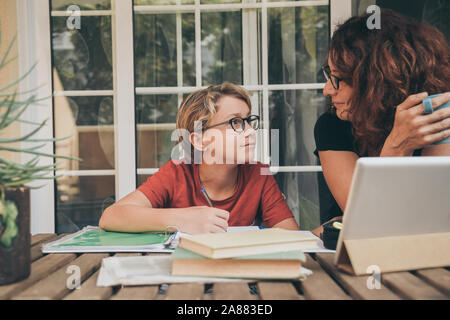 Young student doing homework at home with school books, newspaper and digital pad near his mother. Happy boy looking mum while writing on the copybook Stock Photo