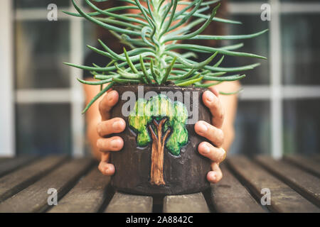 Close up view of hands holding a vase decorated with a tree on a wooden table. Small green plant in a natural ecological pot of recyclable material. Stock Photo