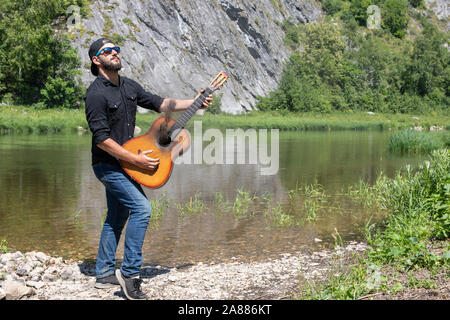young hipster bearded guy, man in a cap plays the guitar with lowered strings against the backdrop of a bright summer landscape Stock Photo