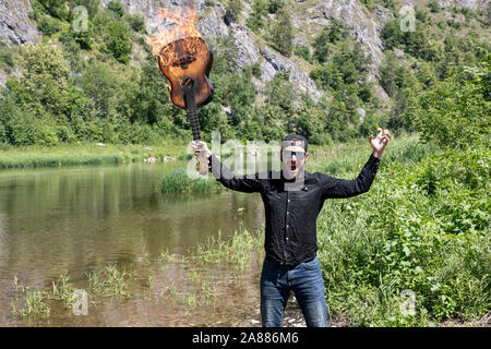 young bearded guy in sunglasses in a black shirt and cap. holding an ignited guitar in raised hands. crazy musician, guitarist on vacation Stock Photo