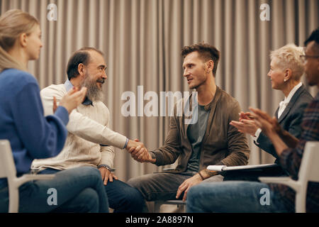 Two men sitting and shaking hands to each other while other people clapping hands they greeting each other Stock Photo