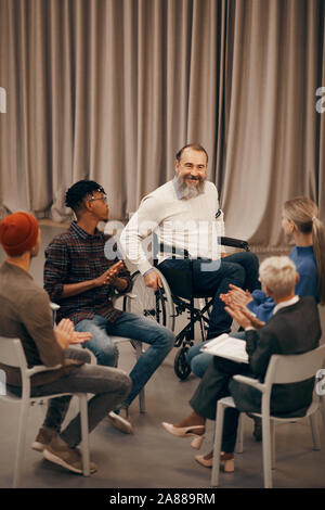 Senior bearded man sitting on wheelchair and smiling while other people clapping hands and greeting him in their class Stock Photo