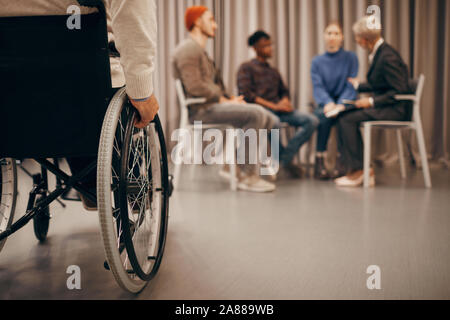 Close-up of disabled man sitting on wheelchair with group of people talking to each other in the background Stock Photo