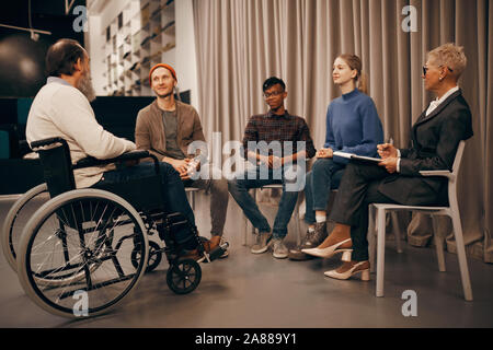 Group of business people sitting on chairs and listening to senior man who sitting in wheelchair at business meeting Stock Photo