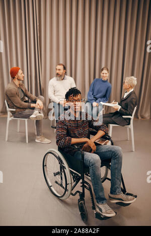 Portrait of African young disabled man sitting in wheelchair and smiling at camera with other people talking in the background Stock Photo