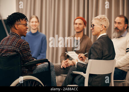 Young African man in wheelchair talking to mature woman while she making notes during therapy lesson Stock Photo