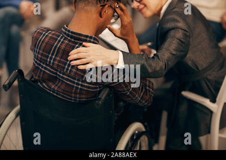 Rear view of African young man sitting in wheelchair and worried about his health with mature woman supporting him Stock Photo