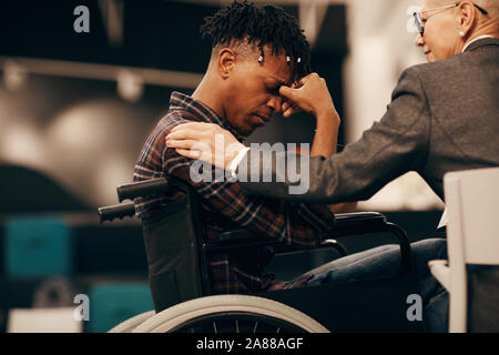 African young disabled man sitting in wheelchair and worried about his disease with woman supporting him Stock Photo