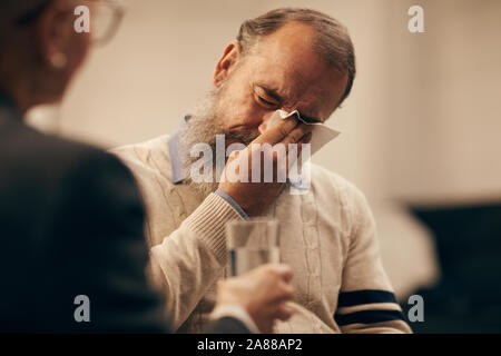 Senior bearded man wiping his tears with napkin with woman giving the glass of water to him Stock Photo