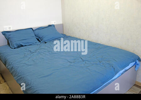 A large gray double bed is in the corner of a light gray room. The bed is covered with monochromatic blue linen, two pillows on the bed and two socket Stock Photo