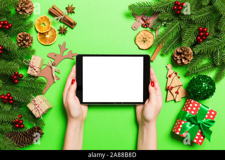 Top view of woman holding tablet in her hands on green background made of Christmas decorations. New Year holiday concept. Mockup. Stock Photo