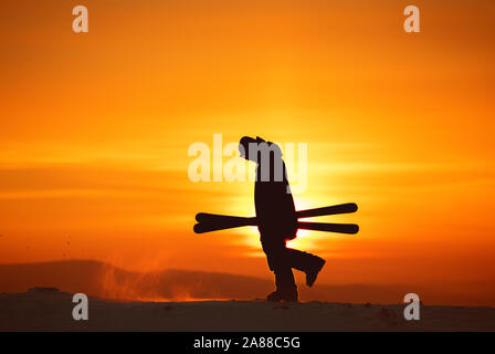 Skiers silhouette goes with ski in hands against sunset sky and sun Stock Photo