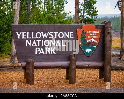 A Yellowstone National Park wooden entrance sign, with fallen autumn leaves underneath and forest trees in the background. Stock Photo