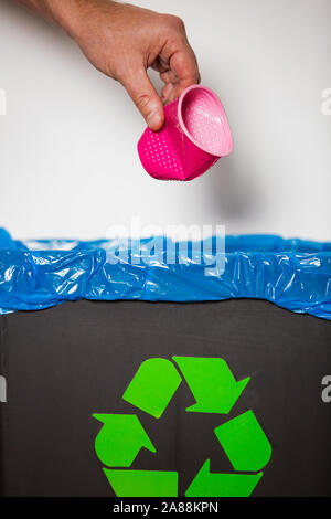Hand putting single-use plastic cup into recycling bin. Person in a house kitchen separating waste. Black trash bin with green bag and recycling Stock Photo