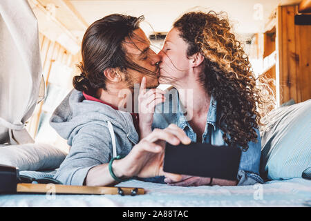 Happy couple kissing in vintage mini van during their roadtrip - Young romantic lovers having tender moments while taking photo with mobile smartphone