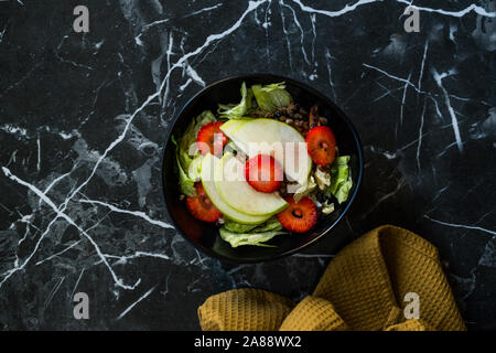 Apple Salad with Strawberry Slices, Buckwheat and Green Leaves in Black Bowl on Dark Granite Surface. Organic Fresh Food. Stock Photo