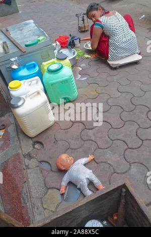 A young woman cutting vegetables on a sidewalk in Mumbai, India, looking over her shoulder at a children's doll lying on the ground Stock Photo