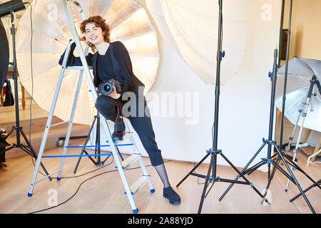 Confident photographer with camera leans against a ladder in the photo studio Stock Photo