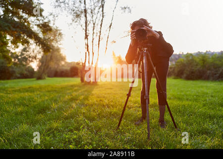 Young woman as landscape photographer with camera on the tripod in the nature while photographing Stock Photo