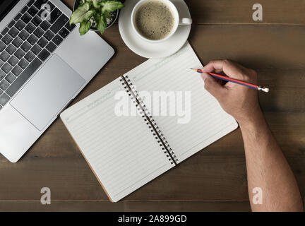 Businessman writing on blank notebook with hot coffee and laptop on wood table, top view Stock Photo