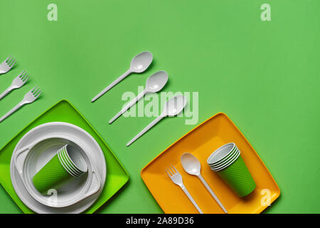 Colorful plastic disposable tableware, such as plates and bowls, four spoons and forks, folded cups on green background with copy space. The concept o Stock Photo