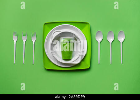 Colorful plastic disposable tableware, such as plates and bowls, three spoons and forks, folded cups on green background with copy space. The concept Stock Photo