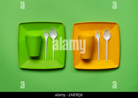 Colored plastic disposable tableware, such as two plates, spoons, forks, folded cups on green background with copy space. The concept of picnic utensi Stock Photo