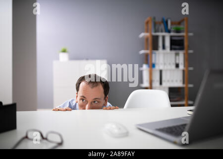 Frightened Young Businessman Hiding Behind Chair At Workplace Stock Photo