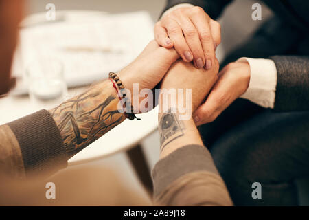 Close-up of mature woman holding hands of young man and supporting him during his visit to psychologist Stock Photo