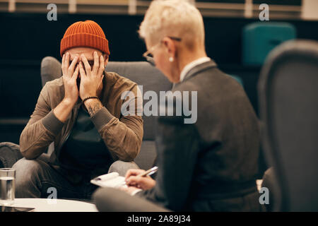 Young worried man sitting on sofa and talking to the mature woman while she making notes at office Stock Photo