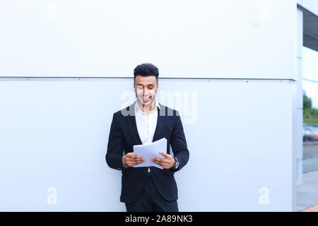 Arabian handsome student smiling, holds and looks at documents near wall of business center. Stock Photo