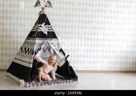 Happy little girl wearing dress sitting near children tent in empty playroom