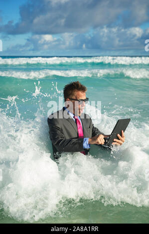 Shocked young businessman in a full suit having a rough time using his tablet computer to surf the web in crashing waves on the shore of the beach Stock Photo