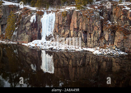 A white frozen waterfall spills down a dark pink-brown sheer cliff of rock reflected in a dark placid river Stock Photo
