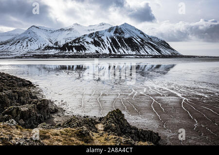 A dramatic snow-covered mountain range rises from the ocean at low tide with rivulets etched into the mud of the seafloor along a rocky coastline Stock Photo