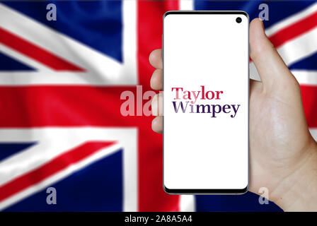 Logo of public company Taylor Wimpey displayed on a smartphone. Flag of UK background. Credit: PIXDUCE Stock Photo
