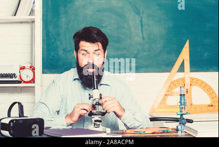 School teacher looking microscope. Fascinating research. Set up microscope. Teacher sit desk with microscope. Man bearded hipster classroom chalkboard background busy with biological observation. Stock Photo