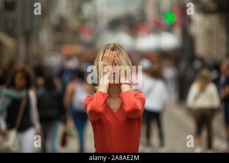 Panic attack in public place.  Woman covers his eyes with his hands surrounded by people walking in crowded street. Stock Photo