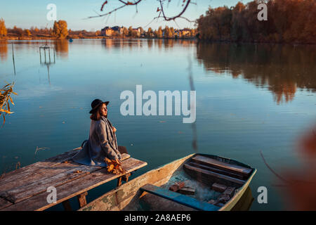 Woman sitting on lake pier by boat admiring autumn landscape and relaxing. Fall season activities