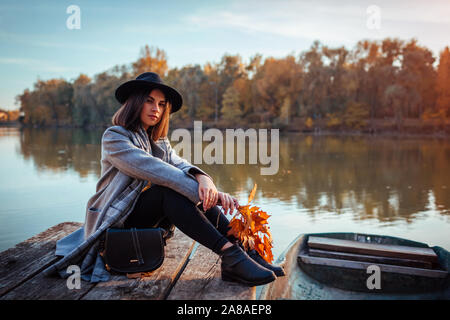 Woman sitting on lake pier by boat admiring autumn landscape and relaxing. Fall season activities Stock Photo