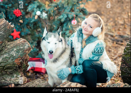beautiful blonde girl sitting in winter park and looking at siberian husky near Christmas tree outdoors. Symbol of new year. Happy New Year and Merry Stock Photo