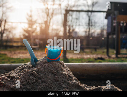 Blue shovel and bucket for children sticking in a mountain of sand on a playground for kids in the summer. In the background is a swing and a slide. Stock Photo