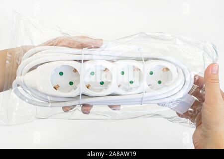 Woman hand holding new four outlets power extension cord in a transparent plastic pakage over white table. Electric power strip for home or office. Stock Photo